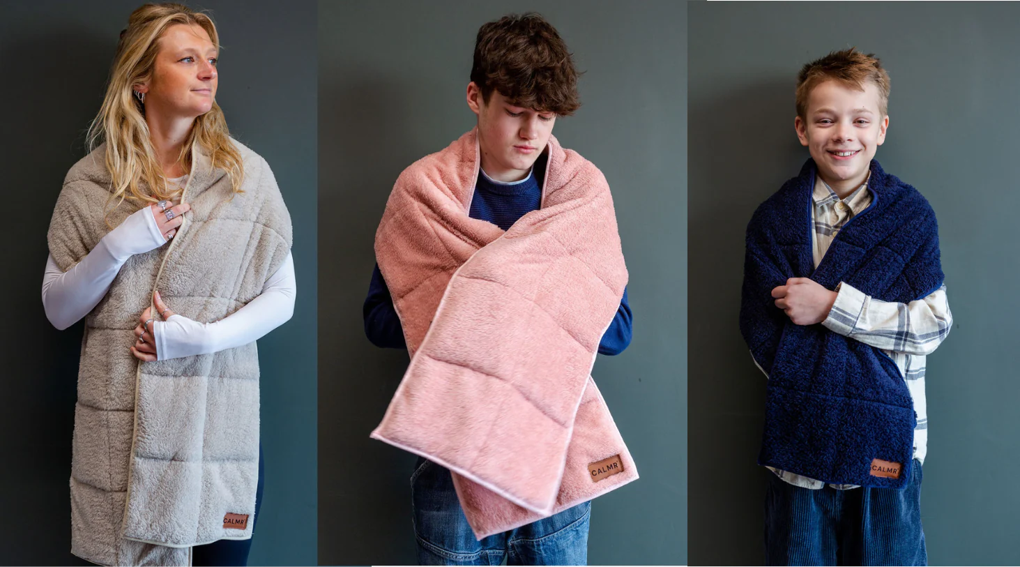 Three people stand against a gray wall, each wearing a different colour of cozy, quilted, CALMR shawls—light gray, pink, and navy blue—smiling or looking content and holding the blanket around their shoulders.